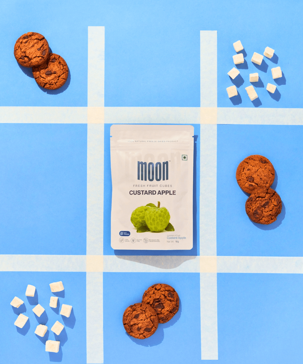 A packet of Moon Freeze Dried Custard Apple + Kiwi snacks surrounded by cookies and sugar cubes arranged on a blue background with white stripes. (Brand Name: MOONFREEZE FOODS PRIVATE LIMITED)