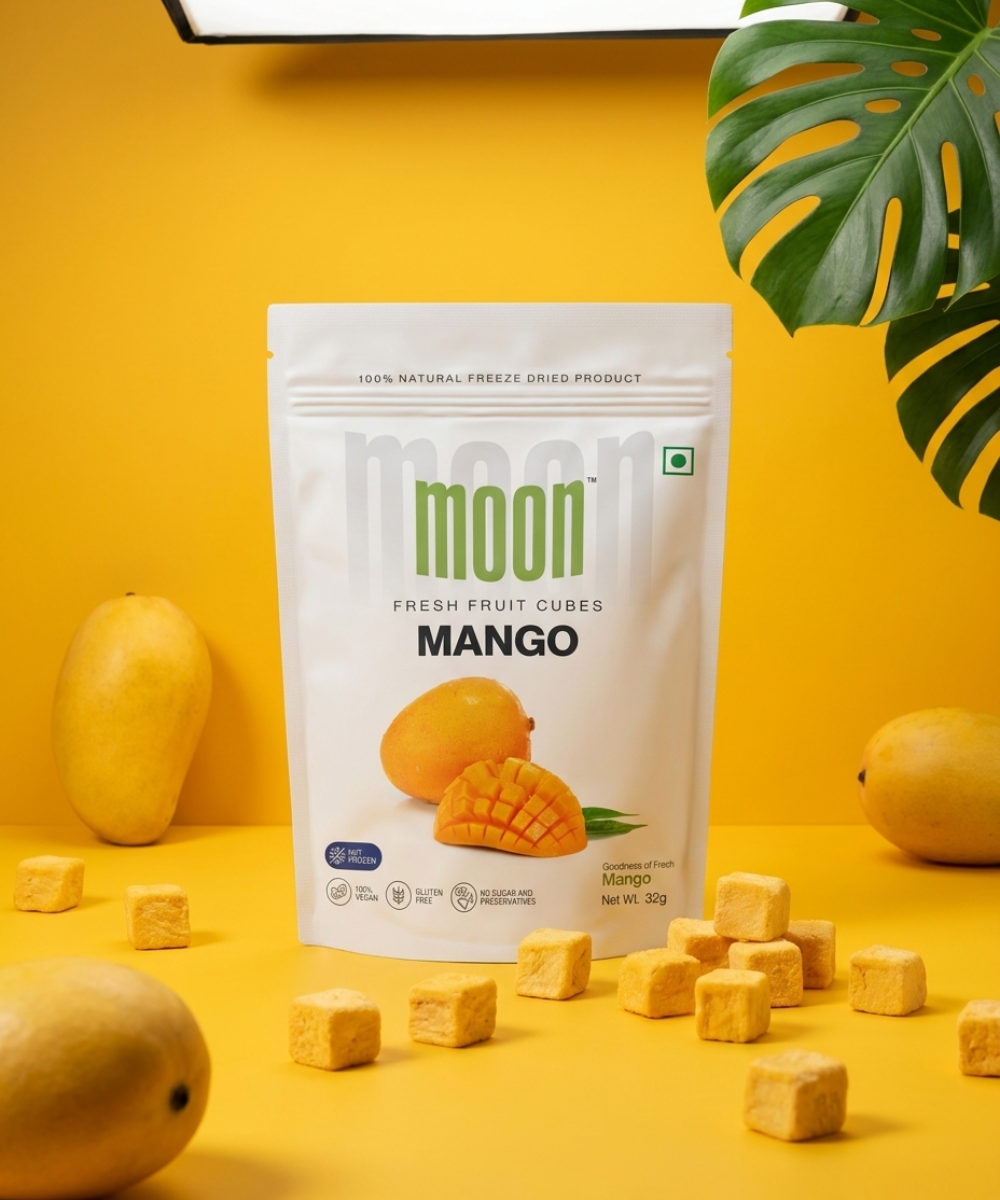 A white packet of Themoonstoreindia’s Moon Freeze Dried Mango Cubes stands upright on a yellow surface with fresh mangoes and cubes around it—a delicious, healthy snack with no added sugar.
