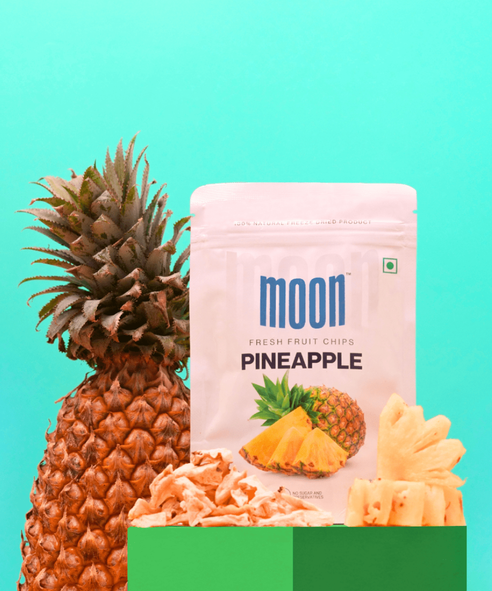 A whole pineapple next to a package of MOONFREEZE FOODS PRIVATE LIMITED's Moon Freeze Moonlight Festival Packs - Summer Edition pineapple fresh fruit chips against a turquoise background.