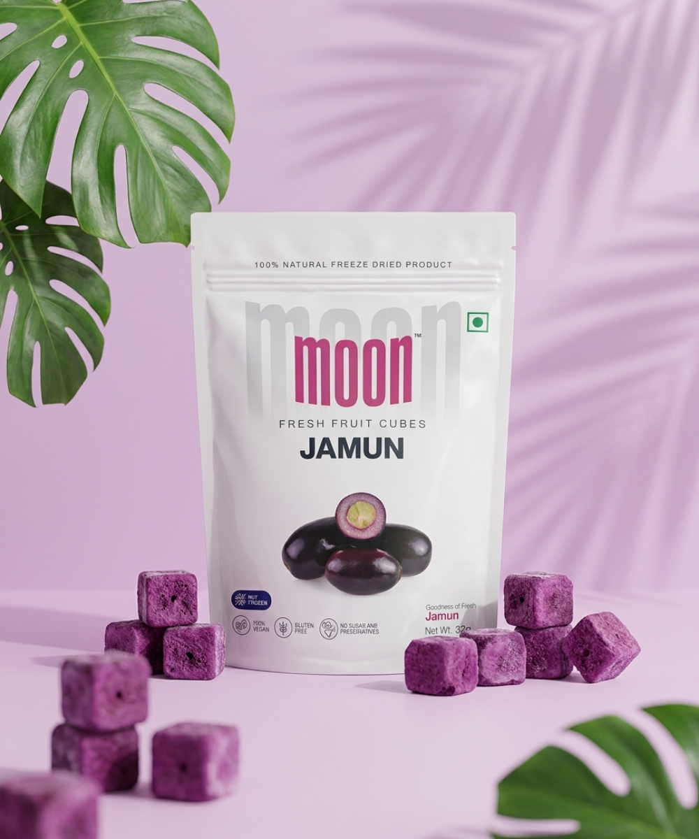 A white packet of "Moon Freeze Dried Jamun Cubes" by Themoonstoreindia stands upright with freeze-dried jamun chips scattered nearby, set against a pink background with green leaves—a tasty, gluten-free treat for natural sugar cravings.