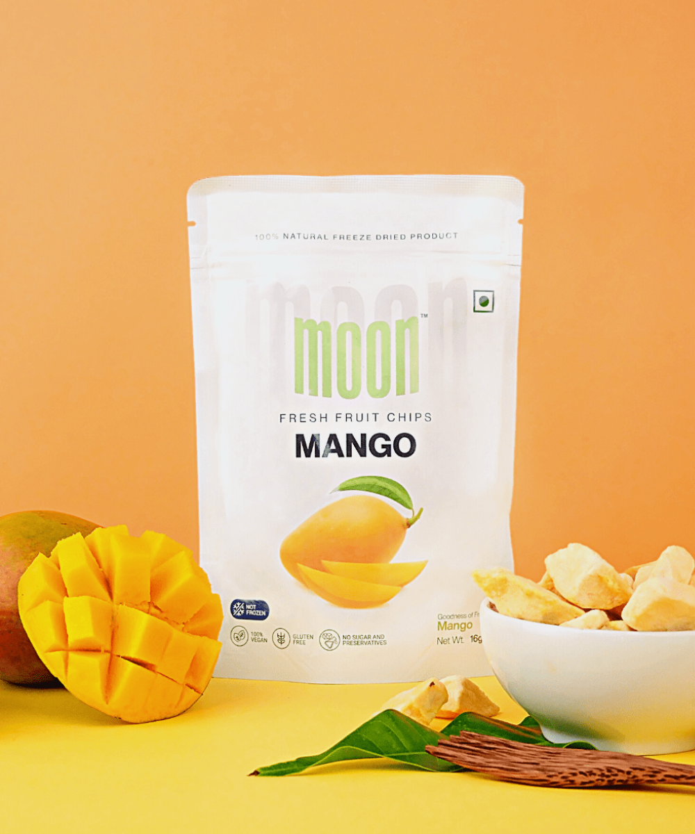 A package of Moon Freeze Moonlight Festival Packs - Summer Edition mango fresh fruit chips by MOONFREEZE FOODS PRIVATE LIMITED against an orange background with mango slices and leaves nearby.