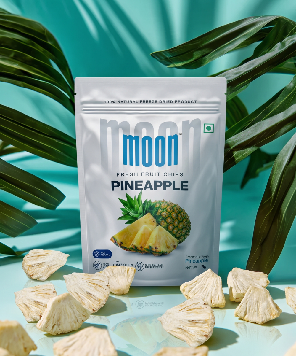 A packet of Moon Freeze Dried Pineapple by Themoonstoreindia stands upright among dried pineapple pieces with tropical leaves and a teal backgroundβan irresistibly healthy snack containing no added sugar.