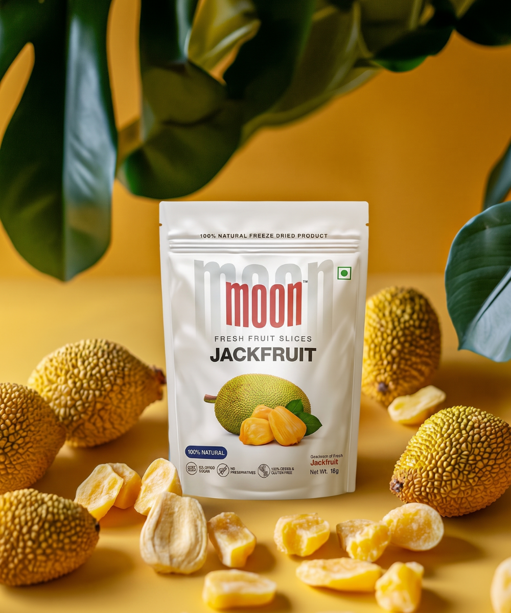 A pack of Themoonstoreindia's Moon Freeze Dried Jackfruit stands upright among fresh jackfruits and loose chips on a yellow surface, with green leaves behind—offering a 100% natural, healthy snack choice.