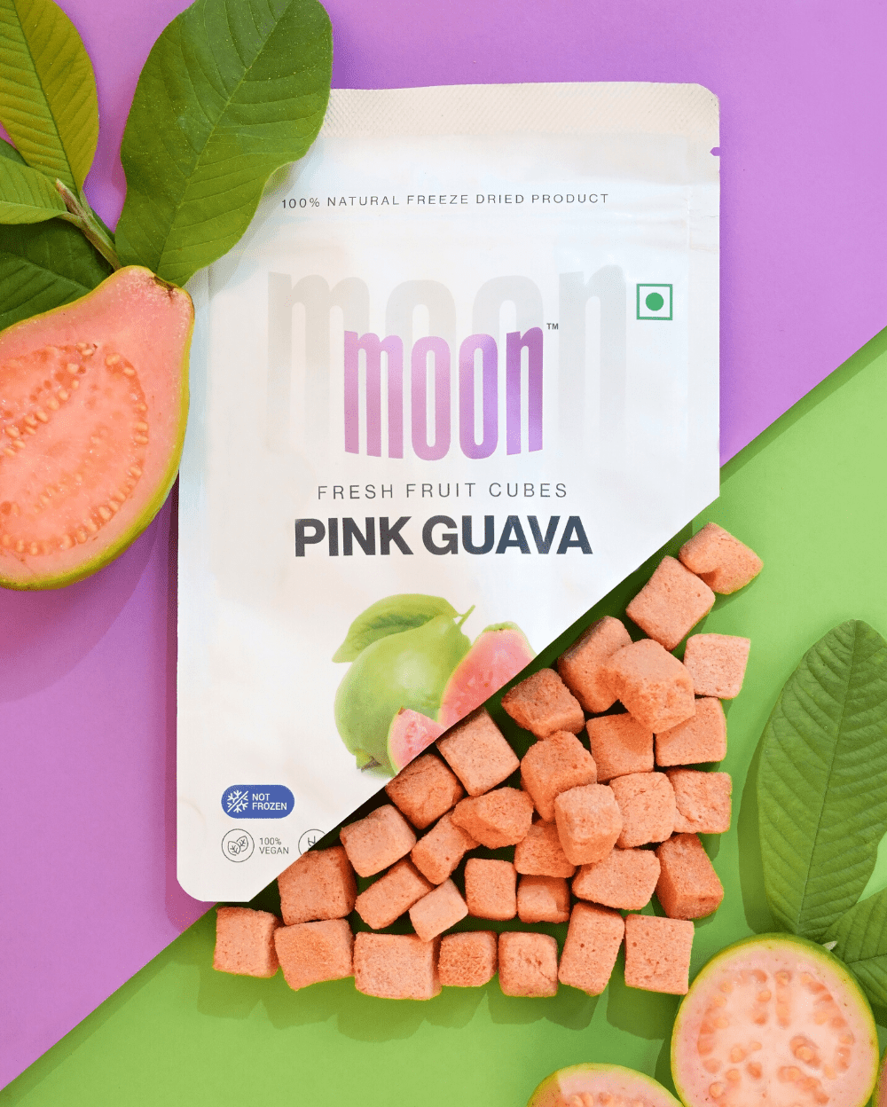 Package of Moon Freeze Dried Pink Guava + Jamun Cubes by MOONFREEZE FOODS PRIVATE LIMITED next to fresh guava slices on a two-tone purple and green background, presenting a captivating option among exotic snacks.