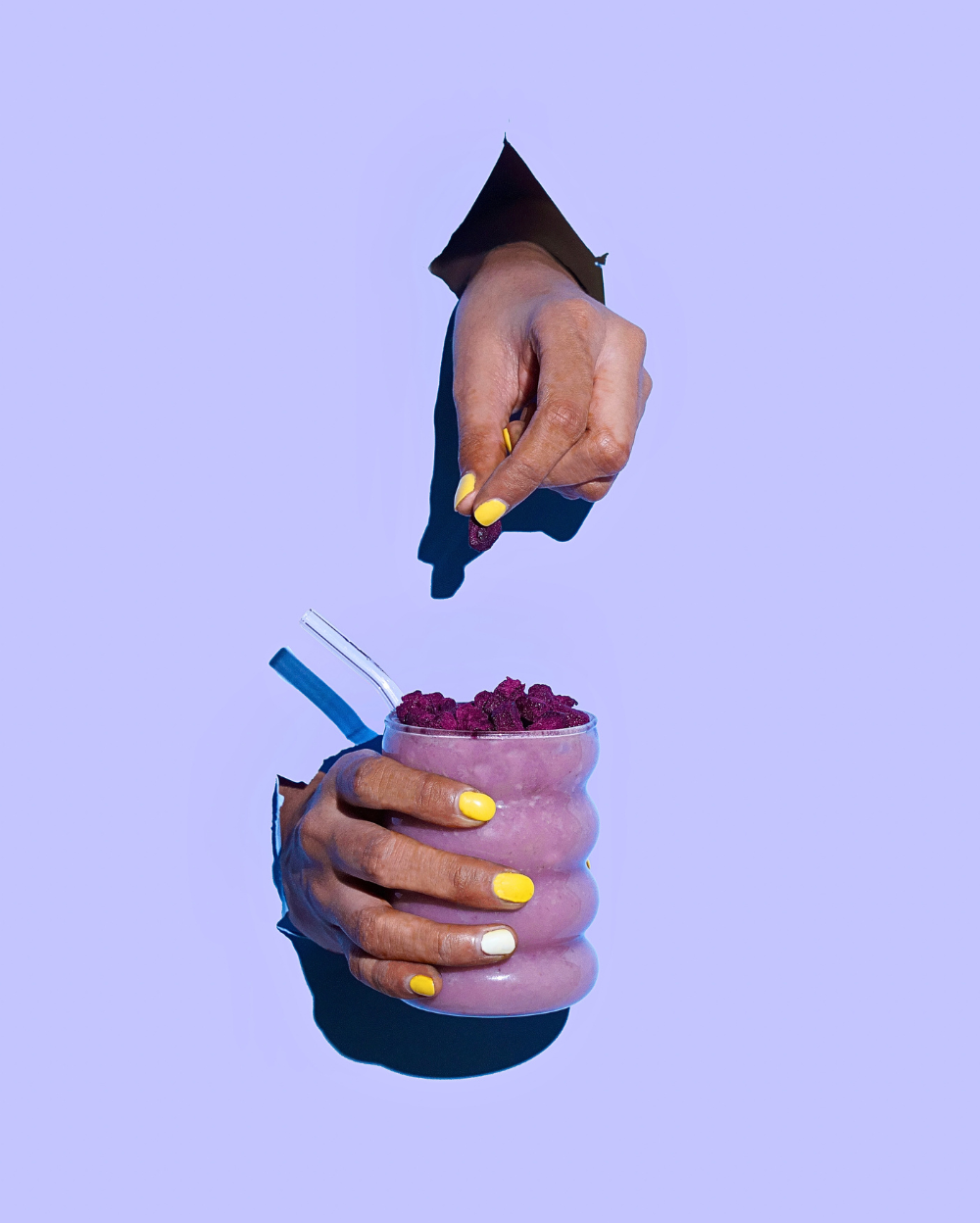 A person with yellow-painted nails holding a glass of pink smoothie topped with Moon Freeze Dried Strawberry + Jamun Cubes against a purple background.
