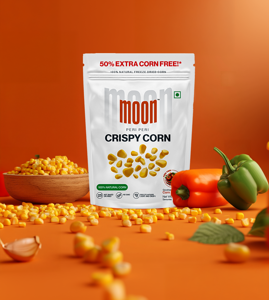 A bag of Freeze Dried Crispy Corn Peri Peri by MOONFREEZE FOODS PRIVATE LIMITED sits among fresh corn, bell peppers, and scattered kernels on an orange backdrop. A delightful guilt-free snack that excites your taste buds with every crunchy bite.