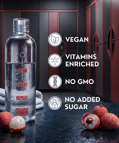 A clear bottle labeled “Lunar Hydration Booster - Lychee Flavour” from MOONFREEZE FOODS PRIVATE LIMITED sits on a gym bench near lychee fruit, displaying icons for vegan, vitamins enriched, no GMO, hydration with electrolytes & no added sugar. Lockers and towel in background.