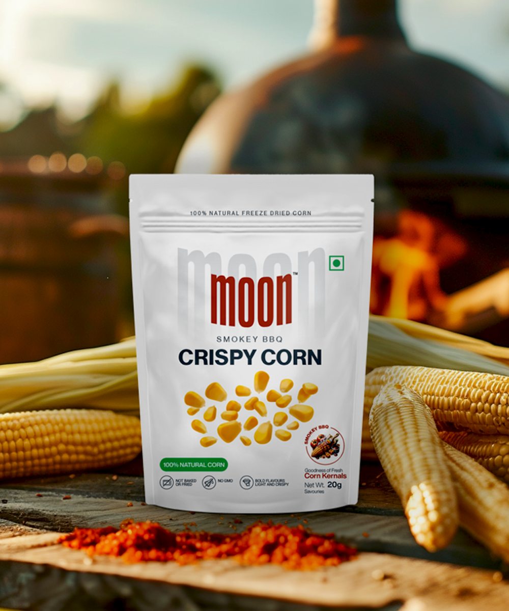 A package of Moon Freeze Orbit Mix Packs - Hydrate & Snack displayed in front of fresh corn cobs and a barbecue grill by MOONFREEZE FOODS PRIVATE LIMITED.