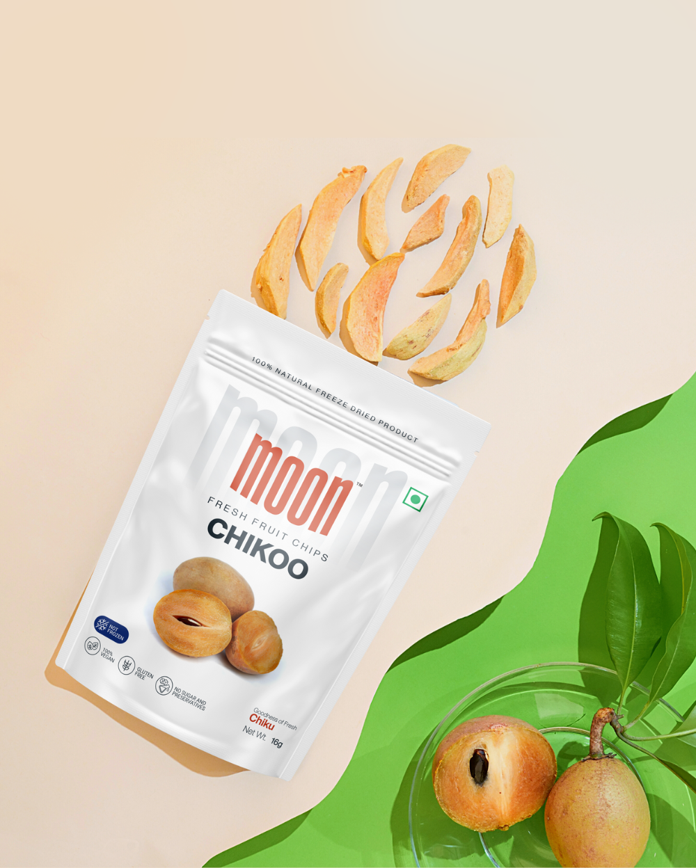 A bag of Crunch & Quench Pack by MOONFREEZE FOODS PRIVATE LIMITED labeled "Chikoo," with scattered chips above. A green branch and chikoo fruit rest on a green surface beside the bag, highlighting the freeze-dried fruits that offer a delicious crunch.