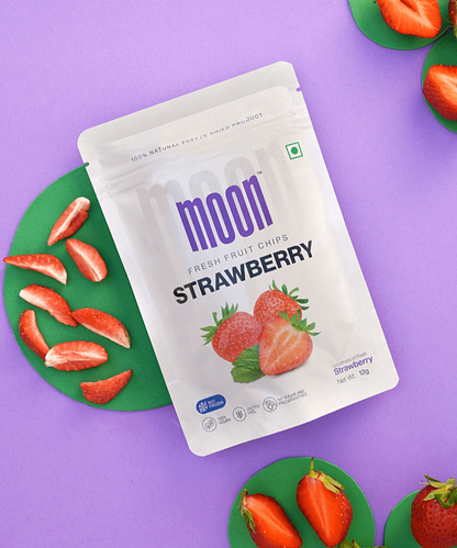 A bag of "Moon Freeze Cosmic Bulk Packs" on a purple surface, surrounded by strawberry halves on green coasters. The packaging shows images of strawberries and product details, highlighting that these freeze-dried fruits offer a celestial snack experience akin to Moon Freeze Cosmic Bulk Packs by MOONFREEZE FOODS PRIVATE LIMITED.