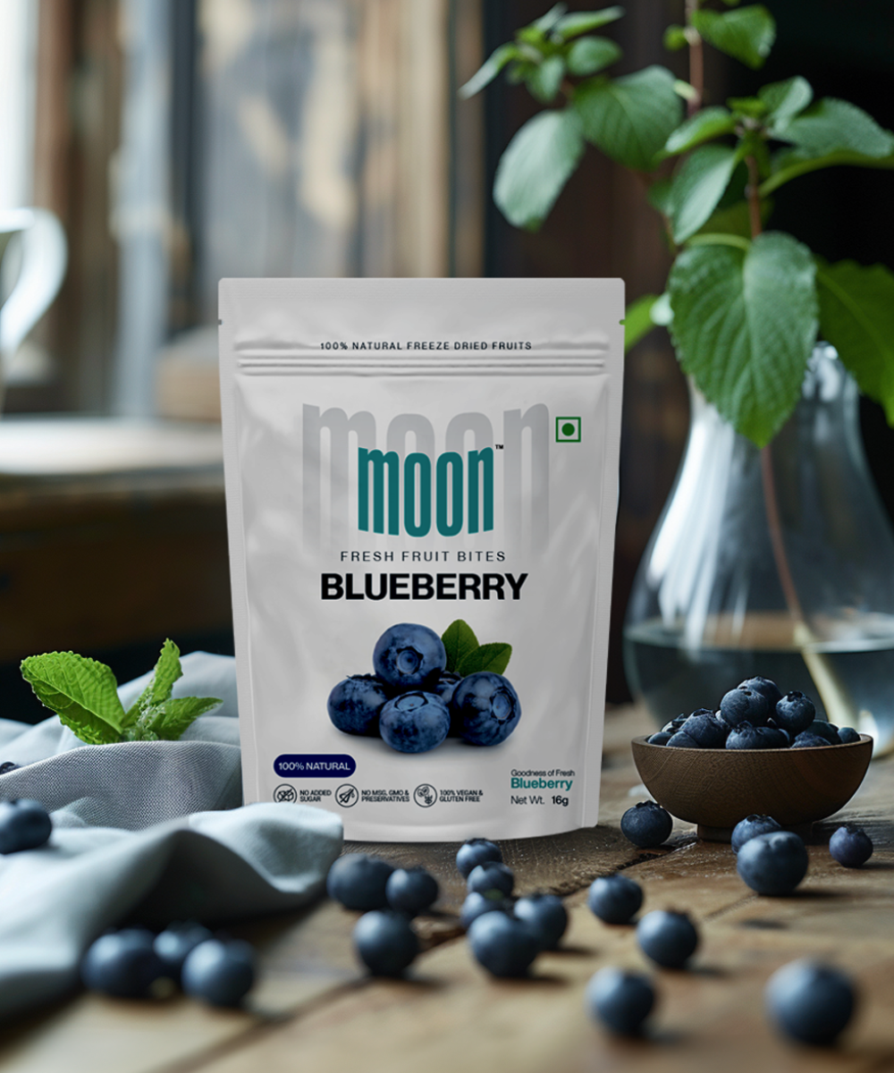 A package of MOONFREEZE FOODS PRIVATE LIMITED's Moon Freeze Dried Strawberry + Blueberry snacks displayed on a table with fresh blueberries and greenery in the background, offering sophisticated snacking options with nutritional benefits.