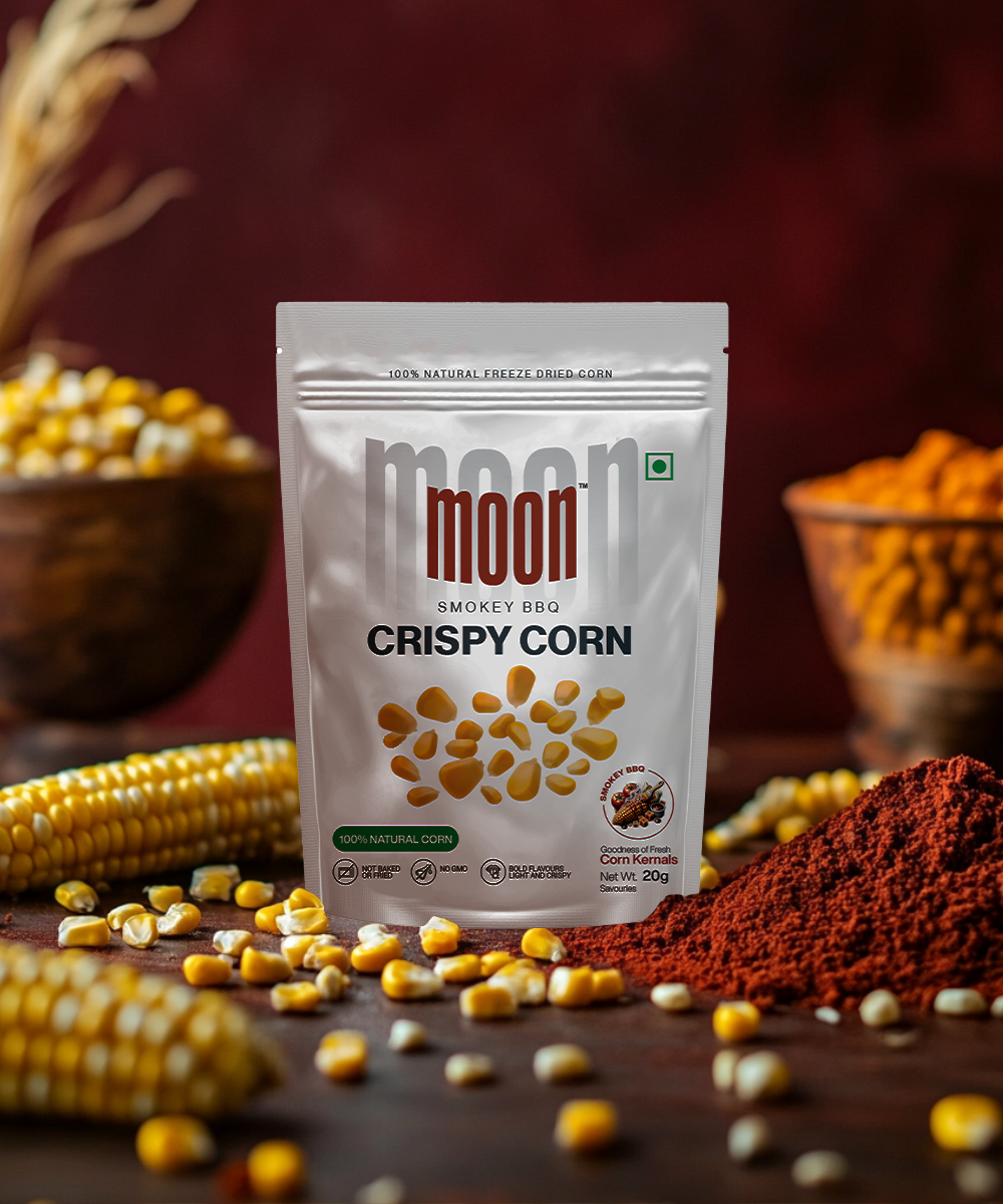 A package of MOONFREEZE FOODS PRIVATE LIMITED's Freeze Dried Crispy Corn Smokey BBQ is showcased on a table, surrounded by scattered plant-based corn kernels, spice powder, and corn cobs in the background.