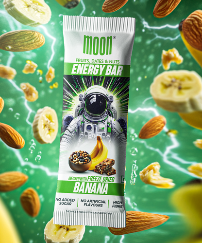 The image showcases a MOON Energy Bar - Banana (6 Pack) from MOONFREEZE FOODS PRIVATE LIMITED, adorned with an astronaut on the wrapper and featuring freeze-dried banana, dates, and nuts. In the background, you'll see floating almond slices, banana chips, and dynamic energy sparks. Made with natural ingredients and high in fiber, it's an ideal meal replacement for those on the go.