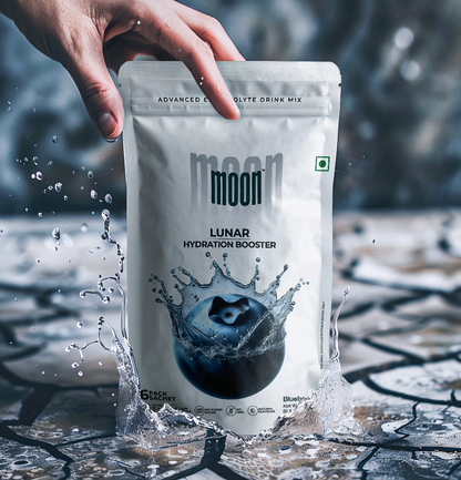 A hand holds the top of a white packet of "Lunar Hydration Booster - Blueberry | Electrolyte Drink Mix" by MOONFREEZE FOODS PRIVATE LIMITED. The drink mix, featuring essential electrolytes and no added sugar, showcases a splash design and is surrounded by splashing water against a textured background.