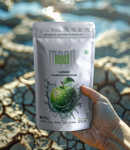 A hand holds a packet of "Lunar Hydration Booster - Green Apple" Electrolyte Drink Mix by MOONFREEZE FOODS PRIVATE LIMITED, featuring a water splash design. This electrolyte-rich drink mix promises superior hydration. In the background, there is blurred outdoor scenery.