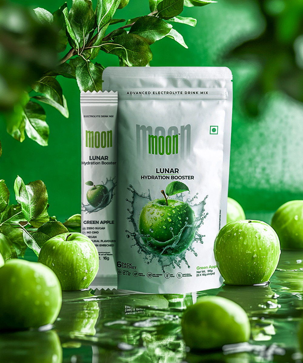 Green Apple flavored hydration booster products from MOONFREEZE FOODS PRIVATE LIMITED, including the Lunar Hydration Booster - Green Apple Electrolyte Drink Mix in a standing pouch and sachet packet, are surrounded by fresh green apples and foliage on a reflective green surface.