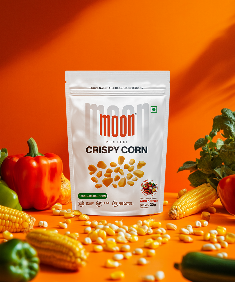 A bag of Freeze Dried Crispy Corn Peri Peri by MOONFREEZE FOODS PRIVATE LIMITED sits among fresh corn, bell peppers, and scattered kernels on an orange backdrop. A delightful guilt-free snack that excites your taste buds with every crunchy bite.