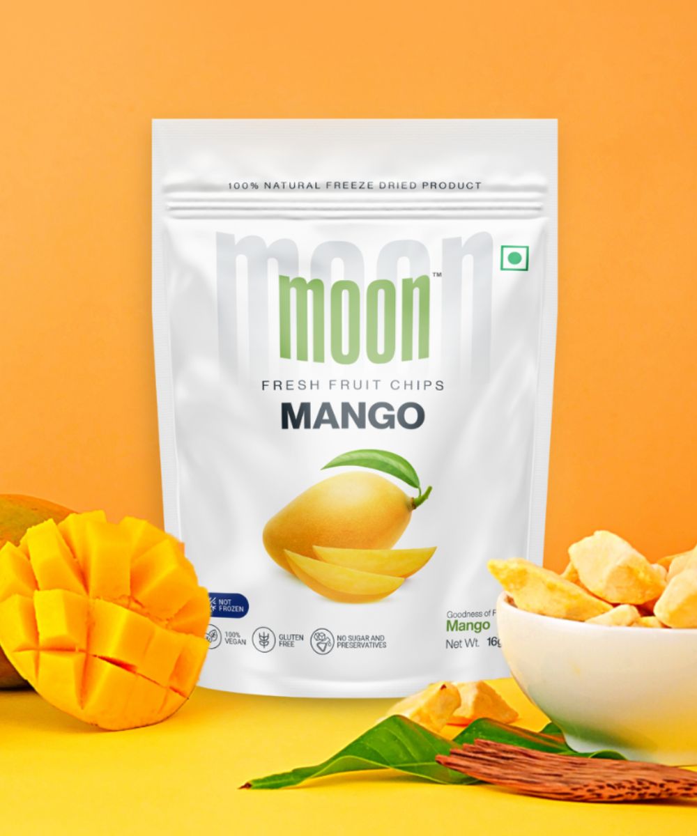 A package of Moon Freeze Dried Mango Chips (16 grams) from Themoonstoreindia is displayed on an orange background, surrounded by a fresh mango, mango slices, and a bowl of mango chips. Packed with high vitamin content and the antioxidant mangiferin, it's a tasty and nutritious treat.