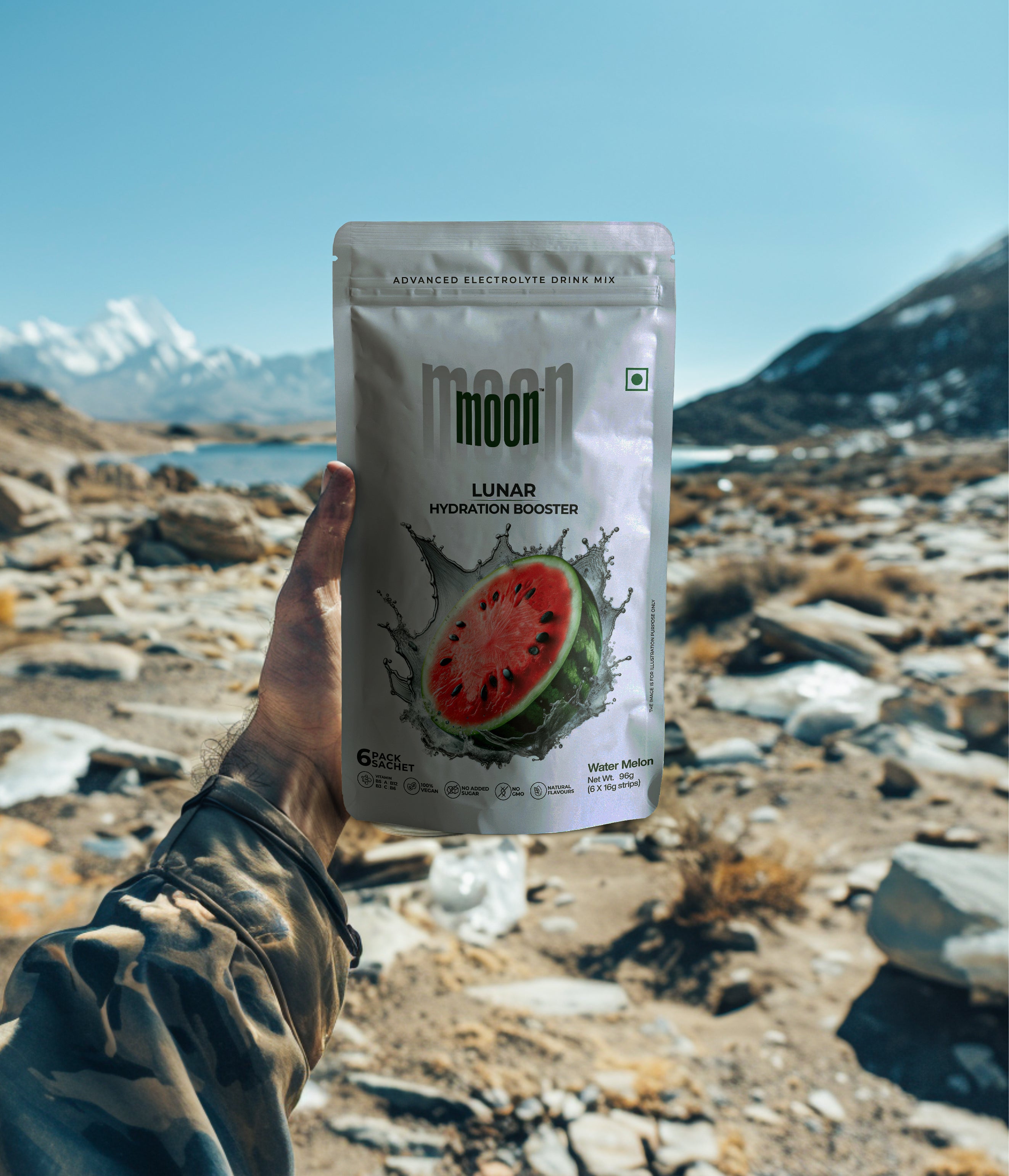 A person holds a packet of "Lunar Hydration Booster - Watermelon" by MOONFREEZE FOODS PRIVATE LIMITED, featuring a watermelon image and a mountainous backdrop. Enhanced with electrolytes and B-vitamin complex, it ensures optimal hydration under the stars.