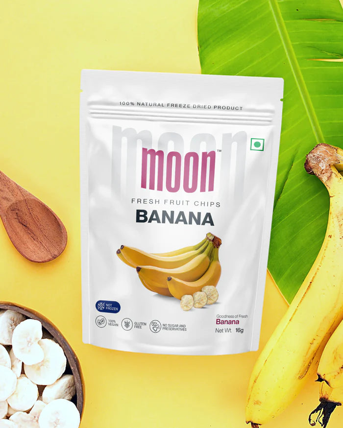 Package of Moon Freeze Dried Banana + Pineapple chips from MOONFREEZE FOODS PRIVATE LIMITED, featuring a whole banana, banana slices, and pineapple pieces against a yellow background with a green leaf and wooden spoon.