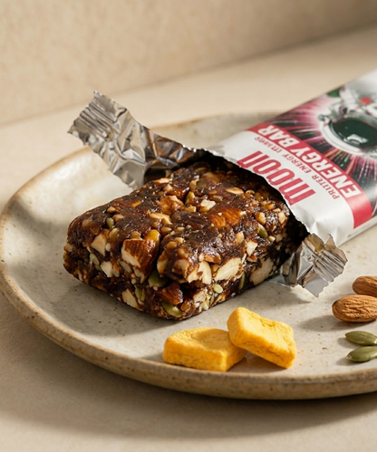 A MOON Energy Bar - Mango by MOONFREEZE FOODS PRIVATE LIMITED sits unwrapped on a plate next to freeze-dried mango pieces and an almond.