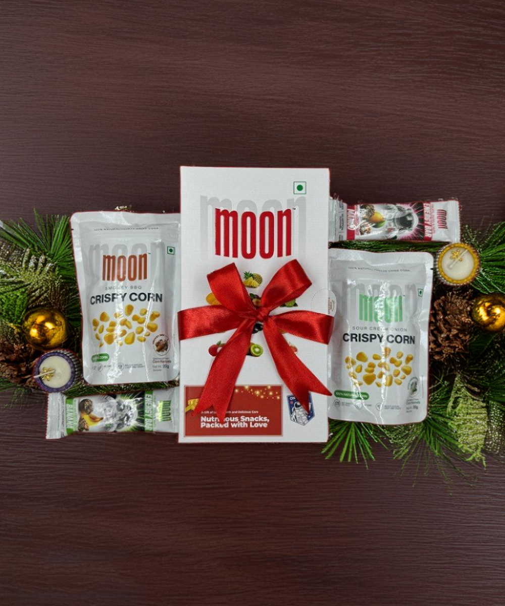 MOONFREEZE FOODS PRIVATE LIMITED's Crisp & Bliss Pack features a gift box adorned with a red ribbon, surrounded by crispy corn snack packs, chocolate, energy bars, and pine decorations on a wooden surface.