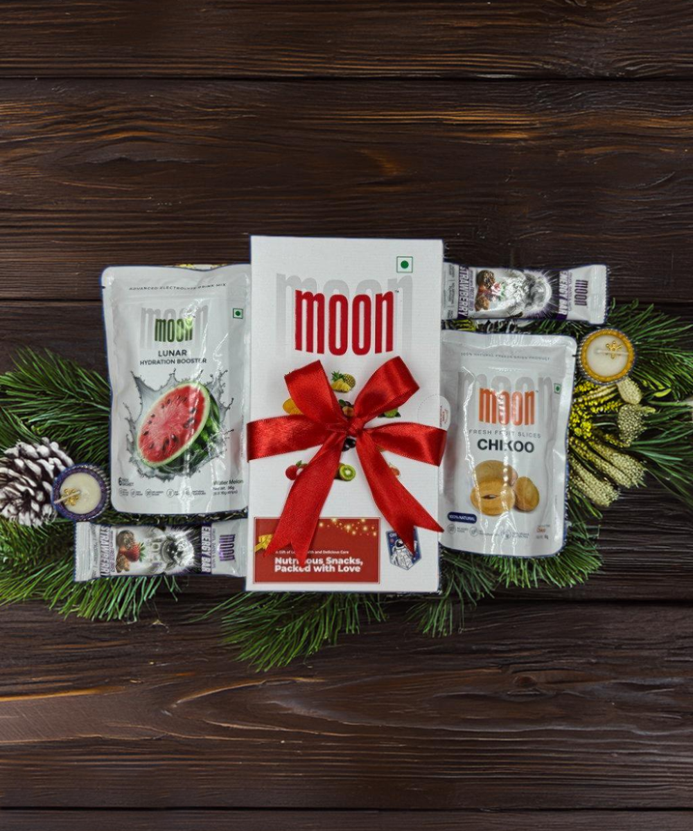 Crunch & Fruity Fun Pack from MOONFREEZE FOODS PRIVATE LIMITED, featuring an assortment of snacks, is beautifully presented in a central box tied with a red ribbon and surrounded by green pine branches and pine cones on a dark wooden background.